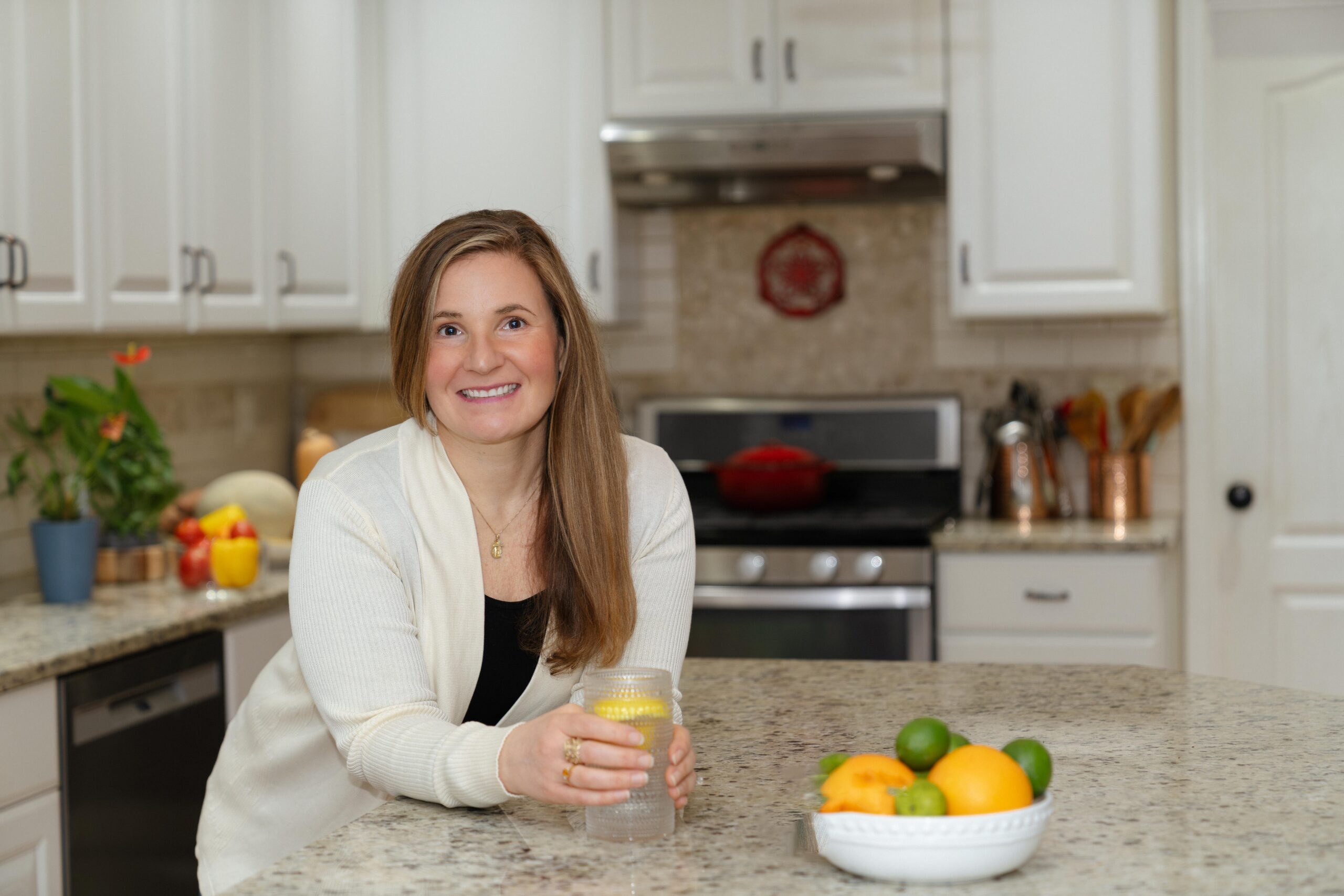 Woman smiling in a bright kitchen, holding a glass with lemon water. Counter has a bowl of fresh fruits; the atmosphere is warm and welcoming.