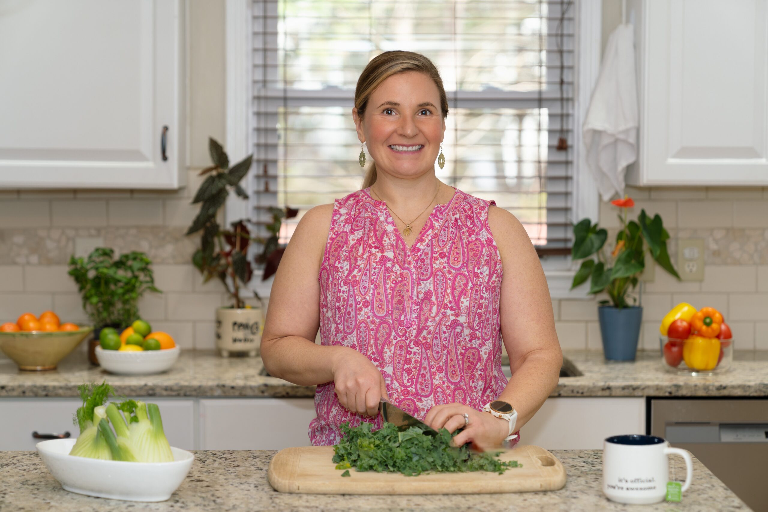 A smiling woman in a pink patterned top chops leafy greens on a wooden board in a bright kitchen. Fresh produce and plants decorate the countertop.