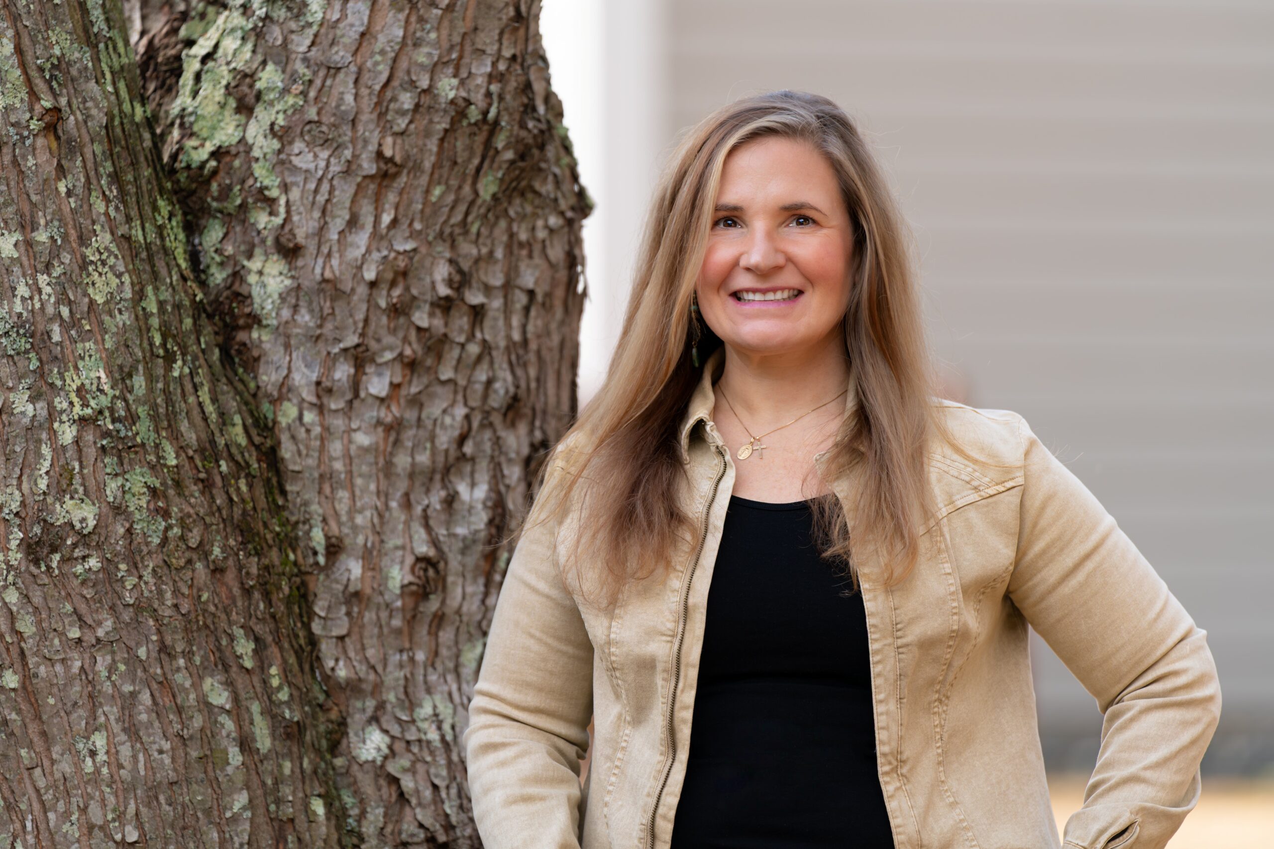 Smiling woman with long brown hair in a beige jacket stands beside a tree with textured bark.
